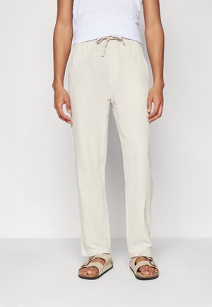 Personne portant un pantalon écru texturé avec cordon de serrage et des sandales beige à bout ouvert avec boucles, debout sur un fond blanc uni.