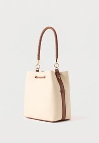 Sac seau beige avec poignées et bandoulière tressées en cuir marron, ferrures dorées et fermeture à cordon sur un fond uni.