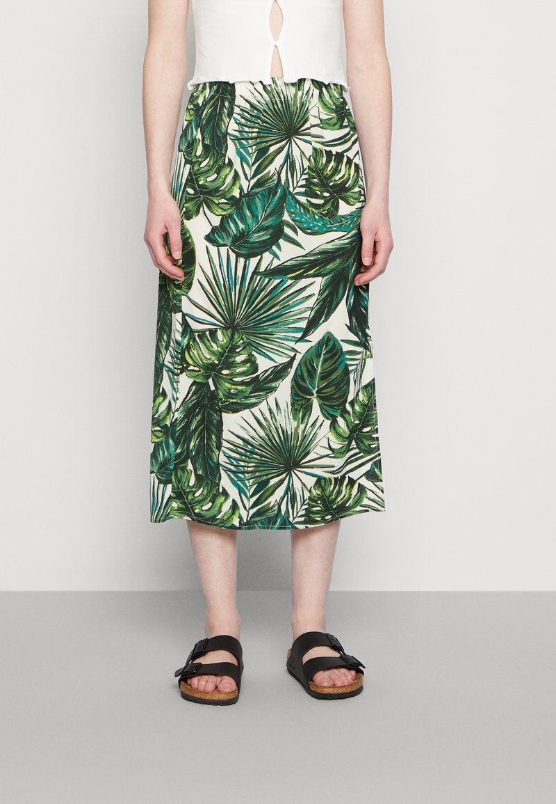 Jupe midi florale avec un imprimé de feuilles tropicales vertes sur un fond clair. Des sandales noires plates avec un détail de boucle sont portées.