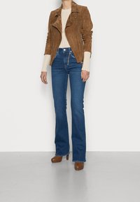 Chaqueta de motorista de ante marrón, suéter de canalé crema, jeans acampanados azul de cintura alta y botas de tacón alto marrones. Accesorios mínimos.