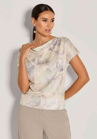 Lichtbeige top met korte mouwen en een bloemenpatroon in zachte pasteltinten. Heeft een gedrapeerde halslijn en een soepele textuur. Gepaard met neutrale broeken.