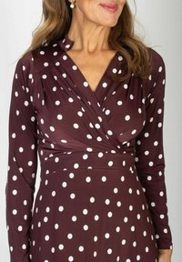 Kobieta w bordowej sukience z marszczeniem w białe kropki i długimi rękawami, widoczna perłowa kolczyk w jednym uchu.