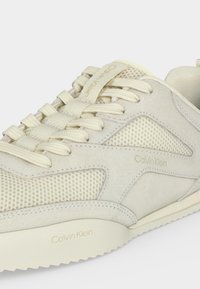 Gros plan d'une basket beige Calvin Klein avec des panneaux en mesh et en daim, une semelle texturée et un logo embossé sur le côté et la semelle.