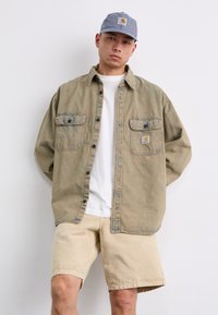 Light brown denim shirt with two front pockets, blue button accents, and a logo patch. Paired with beige shorts and a striped cap.