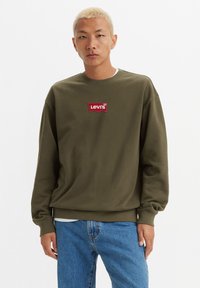 Olivengrønn genser laget av mykt stoff, med et rødt Levi's-logo patch på brystet og elastiske mansjetter. Kombinert med blå jeans.