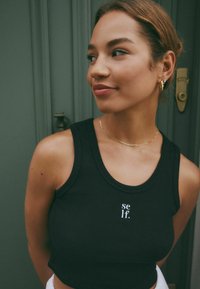 Crni rebrasti crop top s bijelim logotipom 'self.' na prednjoj strani. Model nosi zlatnu ogrlicu i male naušnice na obruče.
