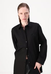 Black button-up shirt with long sleeves, featuring an oval logo patch and a relaxed fit. Made from smooth fabric with a slight sheen.