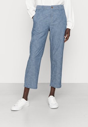 Person wearing light blue straight-leg pants with elastic waist and button, white long-sleeve blouse, and white sneakers, standing on gray floor.