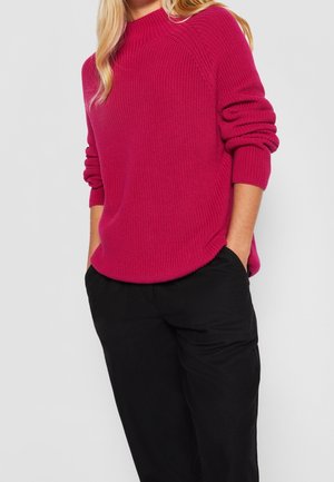 Femme portant un pull en maille rose vif à manches longues et un pantalon noir, les mains dans les poches, sur fond uni.