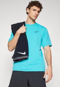 T-shirt Nike turquoise à manches courtes, avec un logo noir. Serviette noire avec un logo blanc drapée sur le bras. Design simple.