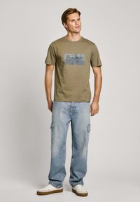 Olijfgroene t-shirt met een grafische print in blauw en grijs, gecombineerd met lichtblauwe cargo-broek en witte sneakers. Eenvoudige ronde halslijn.