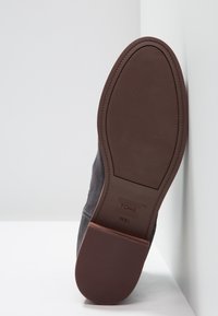 Zapato de ante gris con suela de goma marrón. La suela presenta un patrón de agarre texturizado y un detalle de costura lateral. Se puede ver la marca TOMS.