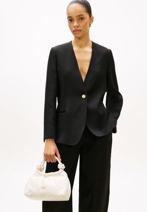 Γυναίκα με μαύρο σακάκι και παντελόνι κρατά λευκή τσάντα χειρός με κόμπους στα χερούλια, στέκεται μπροστά σε απλό ανοιχτό φόντο.