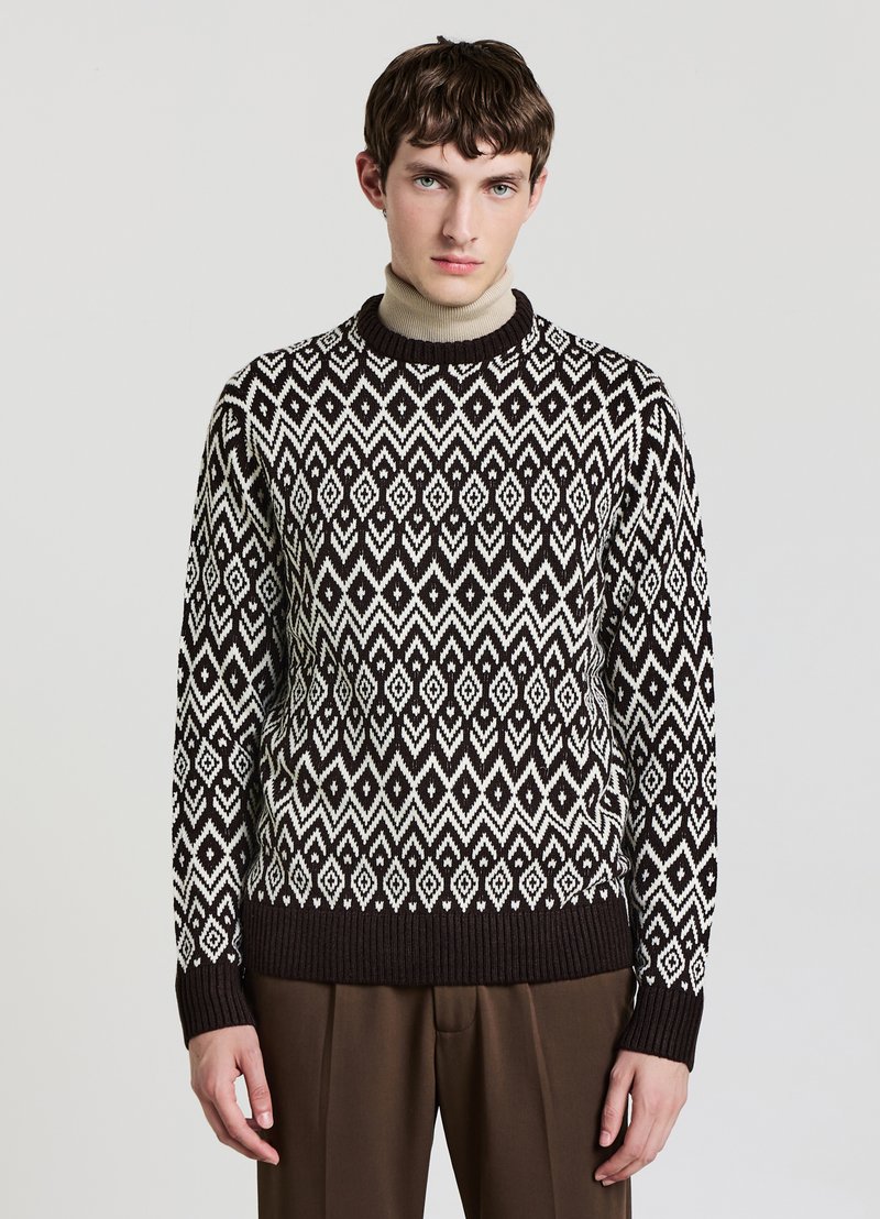 Knit sweater featuring a geometric pattern in black and white, crew neck, and ribbed cuffs and hem, paired with brown trousers.