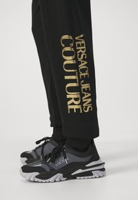 Schwarze Jogginghose mit goldenem "VERSACE JEANS COUTURE" Schriftzug an der Seite; kombiniert mit schwarzen und grauen Sportschuhen mit Mesh-Akzenten.