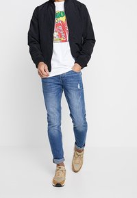 Svart bomberjacka, vit t-shirt med grafik, slitna blå jeans och ljusbruna sneakers. Enkel design med avslappnad passform och lätt material.