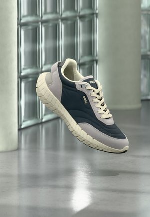 Dunkelblauer und beiger BOSS-Sneaker mit dicker weißer Sohle, Schnürsenkel gebunden, schwebt über poliertem Betonboden nahe einer Glassteinwand.
