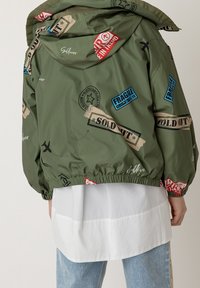 Giacca in nylon verde con grafiche a tema viaggi, dotata di polsini elasticizzati, cappuccio con cordino e dettagli in patch rosso, blu e beige.