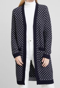 Marinblå stickad cardigan med ett vitt mönster, ribbade ärmar och öppning samt framfickor. Cardigan är lång och har öppen front.