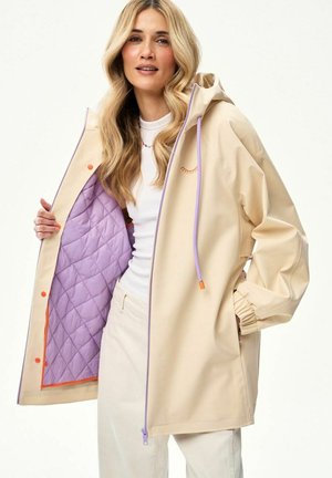 Blondynka w beżowej kurtce z fioletową pikowaną podszewką, fioletowymi troczkami, pomarańczowymi zatrzaskami, białym topem i kremowymi spodniami na białym tle.