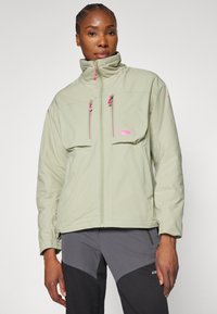 Veste vert clair avec deux poches avant, accents de zip rose, col montant et poignets élastiques. Texture douce, conçue pour une utilisation en extérieur.