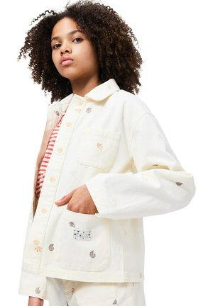 Veste en denim blanc avec motifs de coquillages brodés, fermeture à boutons et poche poitrine ; portée par-dessus un haut rayé rouge et blanc.