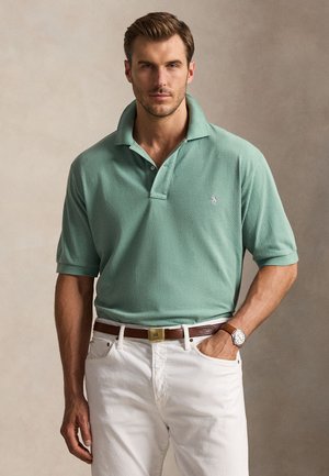 Mand iført en grøn poloshirt med korte ærmer, hvide bukser, brunt bælte og armbåndsur, står med den ene hånd i lommen.