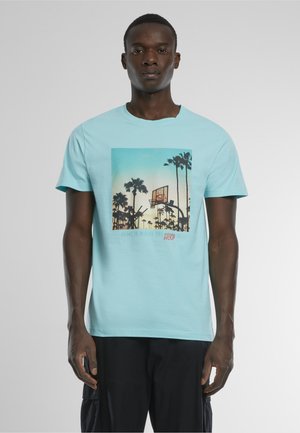 Homme portant un t-shirt bleu clair avec un motif de panier de basket et de palmiers au coucher du soleil, associé à un pantalon noir.