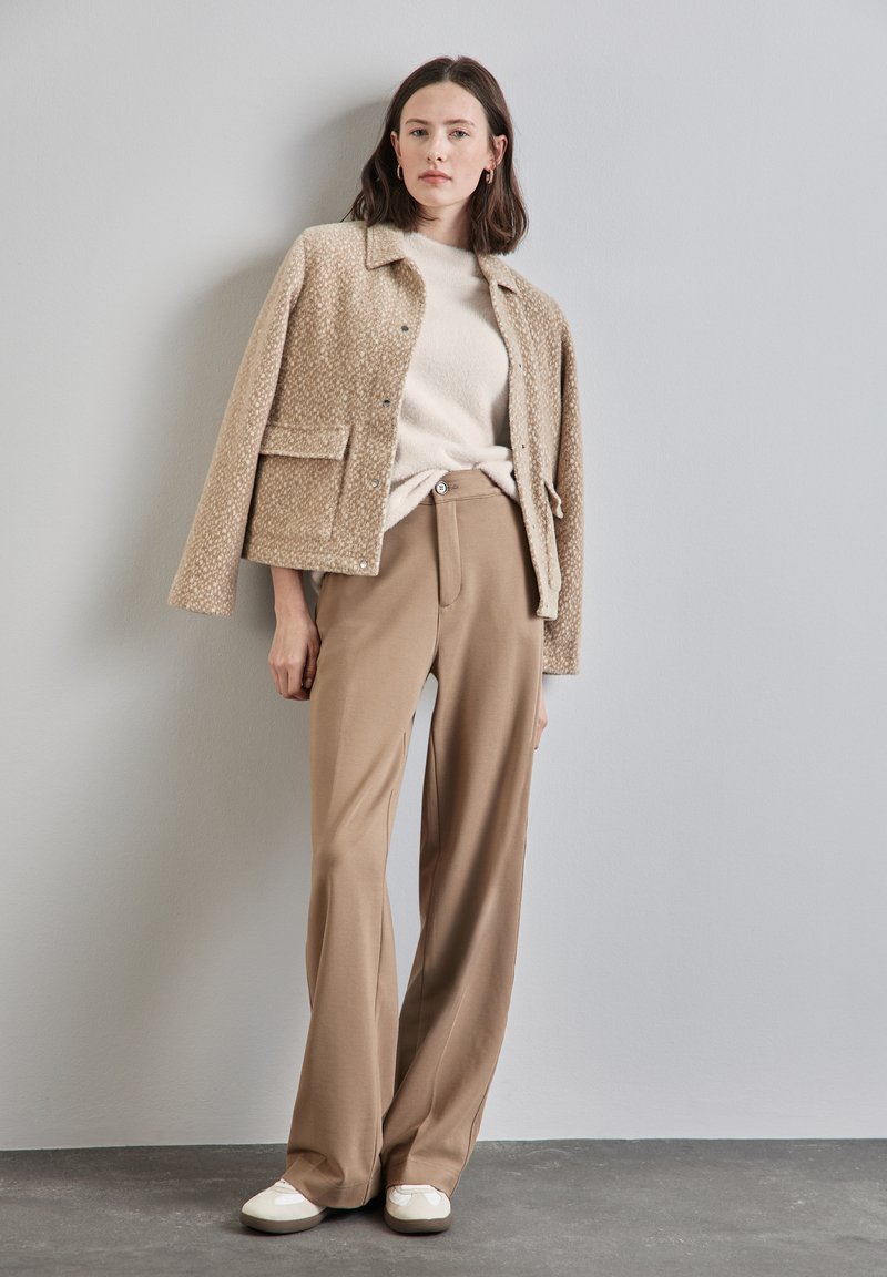 Veste beige texturée avec un col, associée à un pantalon beige clair. L'ensemble comprend des chaussures blanches avec un détail en nœud.