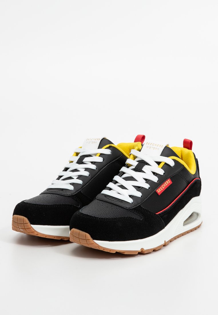 Zwarte suède en mesh sneakers met witte veters, gele accenten en rode branding. Beschikt over een gewatteerde witte zool met een gestructureerde bruine onderkant.