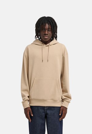 Jong persoon met een donkere huid en dreadlocks, gekleed in een beige geribde hoodie en donkerblauwe spijkerbroek, staand tegen een effen lichte achtergrond.