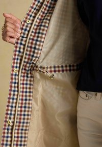 Hand die een opengehouden ruitjesjas toont met beige mesh voering, verstelbare trekkoord en gouden drukknopen.