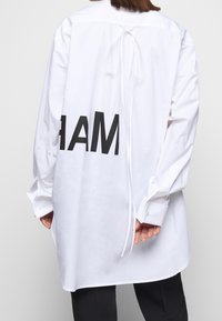 Vit oversized skjorta med knytning bak, prydd med stora svarta bokstäver "AMAR" på baksidan, knäppte ärmslut och en mjuk bomullstextur.