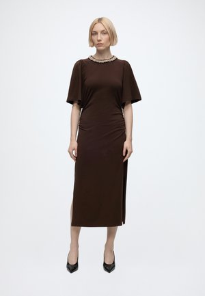 Femme debout, face à l’avant, portant une robe midi marron foncé avec des manches courtes évasées, un col orné, une fente latérale et des talons noirs pointus.