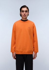 Felpa arancione con collo rotondo e polsini a coste; piccolo logo sul petto. Abbinata a pantaloni neri, il modello si trova davanti a uno sfondo semplice.