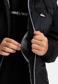 Händer som zippar en svart kviltad väst som bärs över en svart skjorta med en synlig vit Quiksilver-logotyp på bröstet.