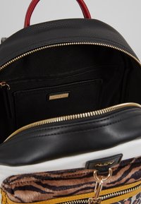 Mochila de estampado animal en blanco, negro y dorado, con cremalleras doradas, asa roja y compartimento principal parcialmente abierto que muestra un bolsillo con cremallera en el interior.