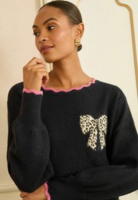Čierny sveter s ružovým volánkovým výstrihom a manžetami, ktorý má na prednej strane detail v tvare leopardieho uzla. Mäkká látka s voľným strihom.