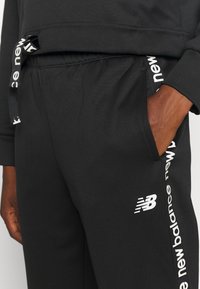 Zwarte joggingbroek met een logo op het linkerbeen, zijzakken en een contrasterende witte trekker met merktext. Zachte textuur.