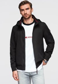Sort letvægtsjakke med lynlås, hætte, elastiske manchetter og røde accenter. Båret over en hvid t-shirt med trykt tekst og jeans.