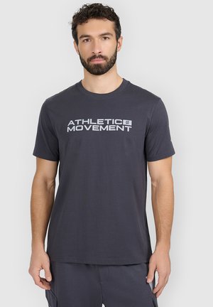 Mężczyzna z brodą w ciemnoszarej koszulce z napisem "Athletic Movement", stojący na jednolitym jasnoszarym tle.