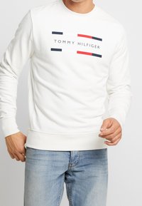 Muž v bílém dlouhém svetru značky Tommy Hilfiger s logem a světlých modrých džínách, stojící na bílém pozadí.