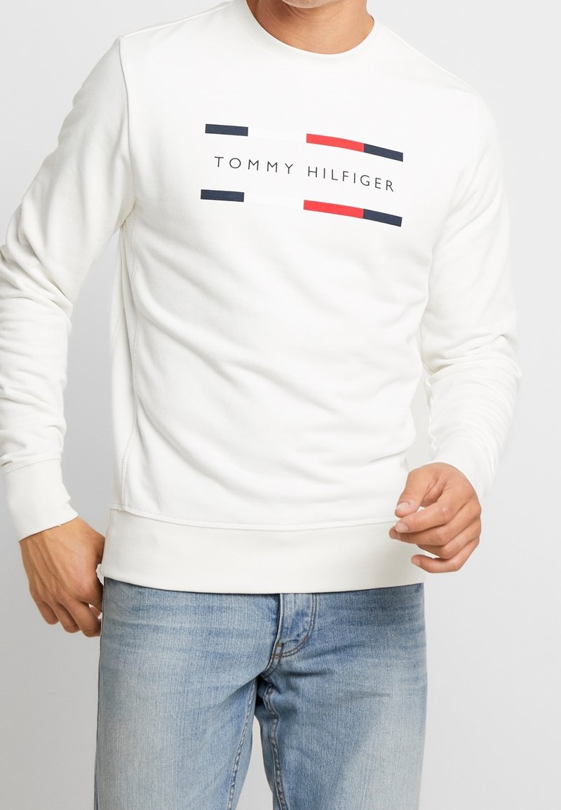 Muž v bílém dlouhém svetru značky Tommy Hilfiger s logem a světlých modrých džínách, stojící na bílém pozadí.