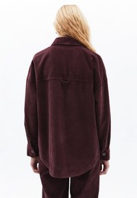 Burgundy kordflorskjorta med en rundad nederkant, knappade ärmslut och en rygghalsdetalj. Tyget har en vertikal ribbad textur genomgående.