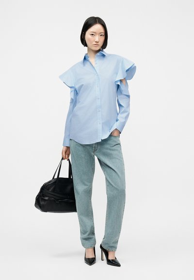 Femme portant une chemise bleu clair à manches à volants, un jean bleu clair, des talons noirs pointus, et tenant un grand sac à main noir sur fond blanc.