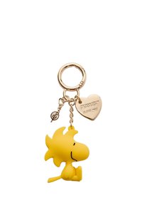 Keltainen kuminen avaimenperä, joka on muotoiltu sarjakuvamaiseksi linnuksi, jolla on hymy, kiinnitetty kultaketjuun ja -renkaaseen, jossa on sydänkoriste, johon on kaiverrettu "PEANUTS".
