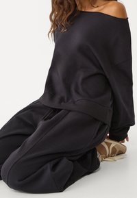 Czarna bluza z opuszczanymi ramionami o luźnym kroju i ściągaczach na rękawach, zestawiona z luźnymi czarnymi spodniami. Gładka faktura, minimalistyczny design.