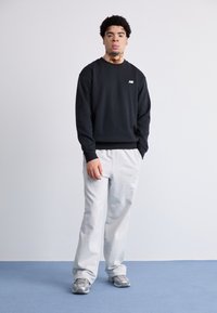 Czarny sweterek z prążkowanymi mankietami, szare dresy i szare buty sportowe. Prosty design z logo New Balance na klatce piersiowej.