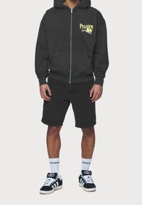 Felpa nera con zip e scritta gialla "PEGADOR", abbinata a pantaloncini in denim neri e sneakers blu scuro. I calzini bianchi riportano il logo "PEGADOR".