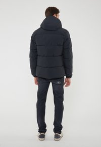 Giacca puffer da uomo di un blu navy scuro con un ampio cappuccio, design trapuntato e polsini elasticizzati. Abbinata a jeans scuri e sneakers casual.